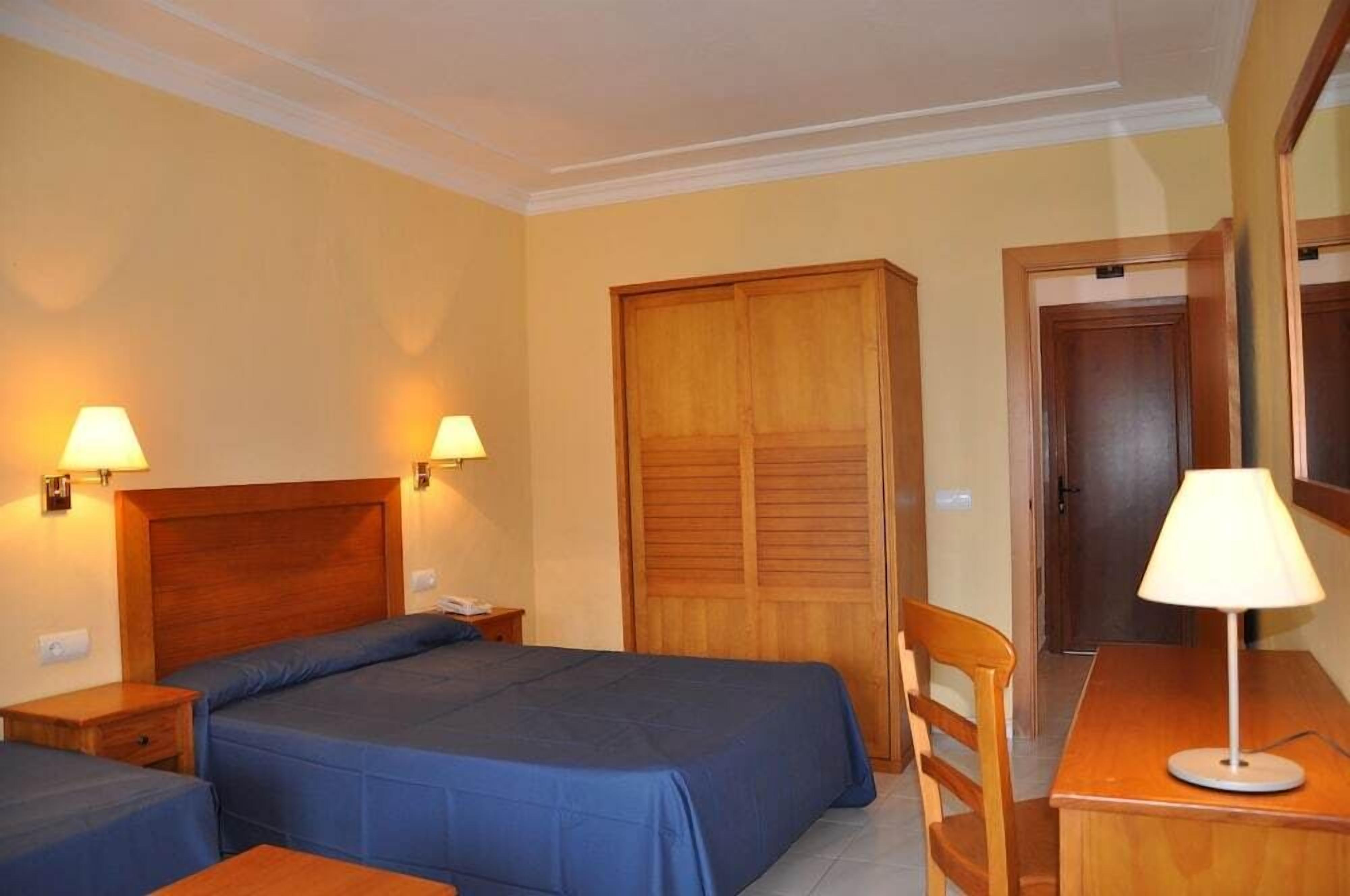 Camera d'albergo con due letti, un'armadio in legno, una scrivania con lampada e una sedia.