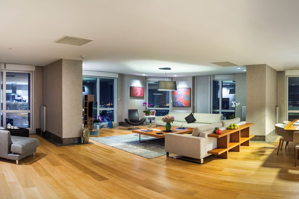 Ein modernes Wohnzimmer mit Sofa, Couchtisch und Essbereich. Es gibt eine Küche mit weißer Arbeitsplatte und Barhocker. Das Zimmer verfügt über große Fenster mit Blick auf die Stadt.