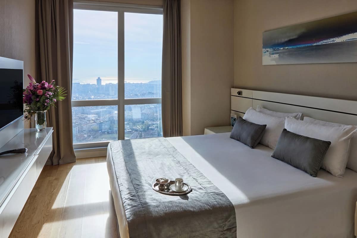 Ein Hotelzimmer mit einem großen Bett, einem Fernseher, einem Blumenstrauß und Blick auf die Stadt durch das Fenster.
