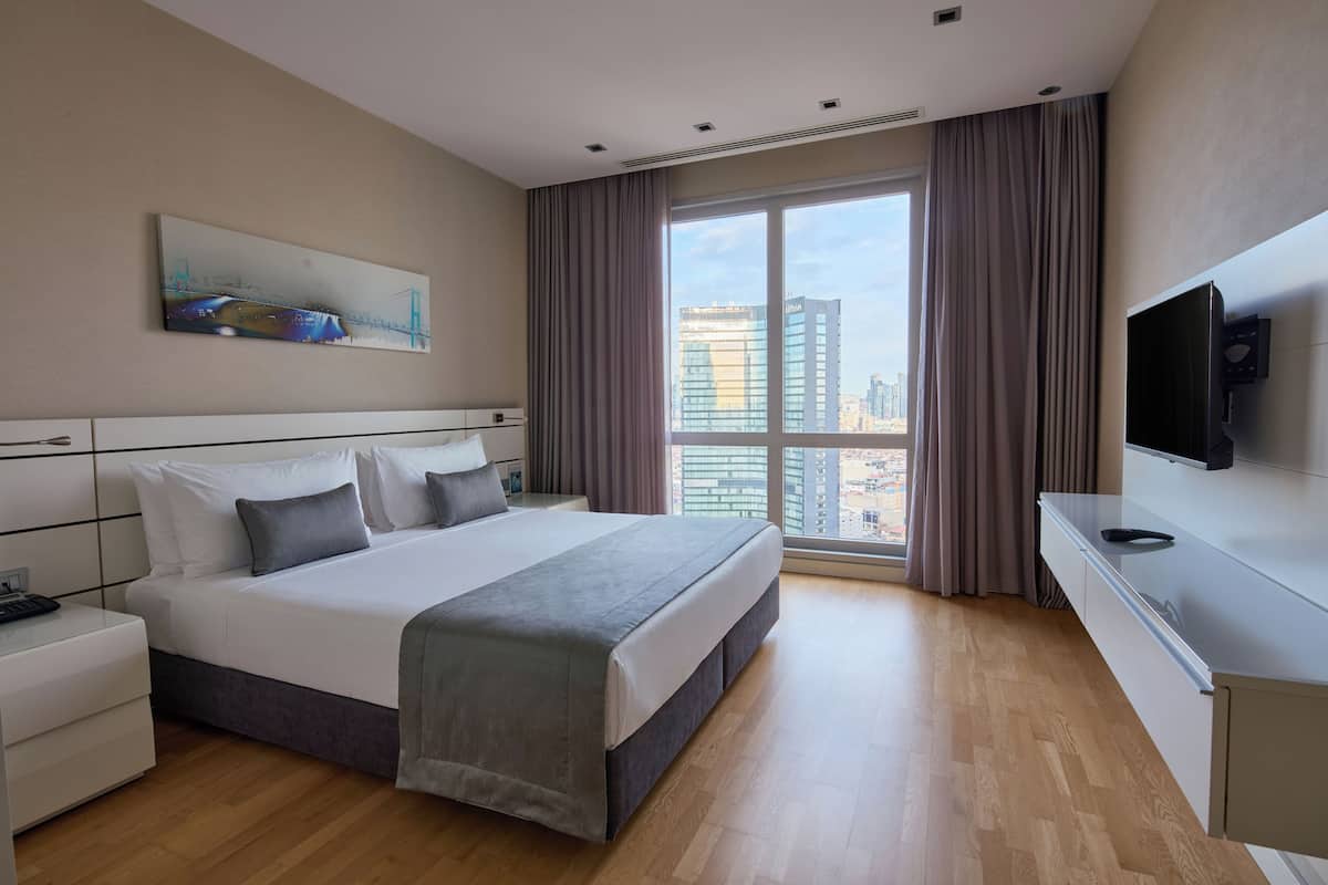 Ein Hotelzimmer mit einem großen Bett, einem Flachbildfernseher, einem Nachttisch und einem Fenster mit Blick auf die Stadt.