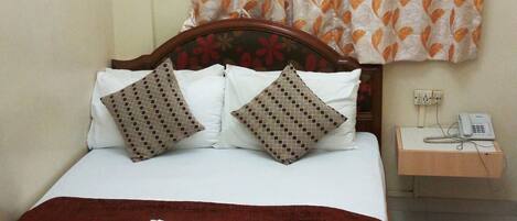 A single bed with a wooden frame, white bedding, patterned pillows, a red blanket, and a floral curtain.