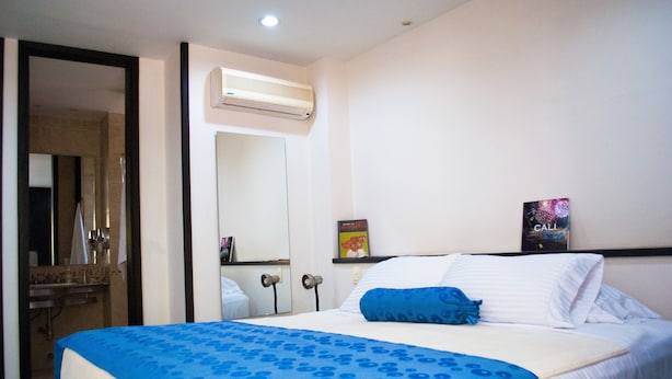 A hotel room with a bed, a mirror, an air conditioning unit, a shelf with a book, and a poster on the wall.
