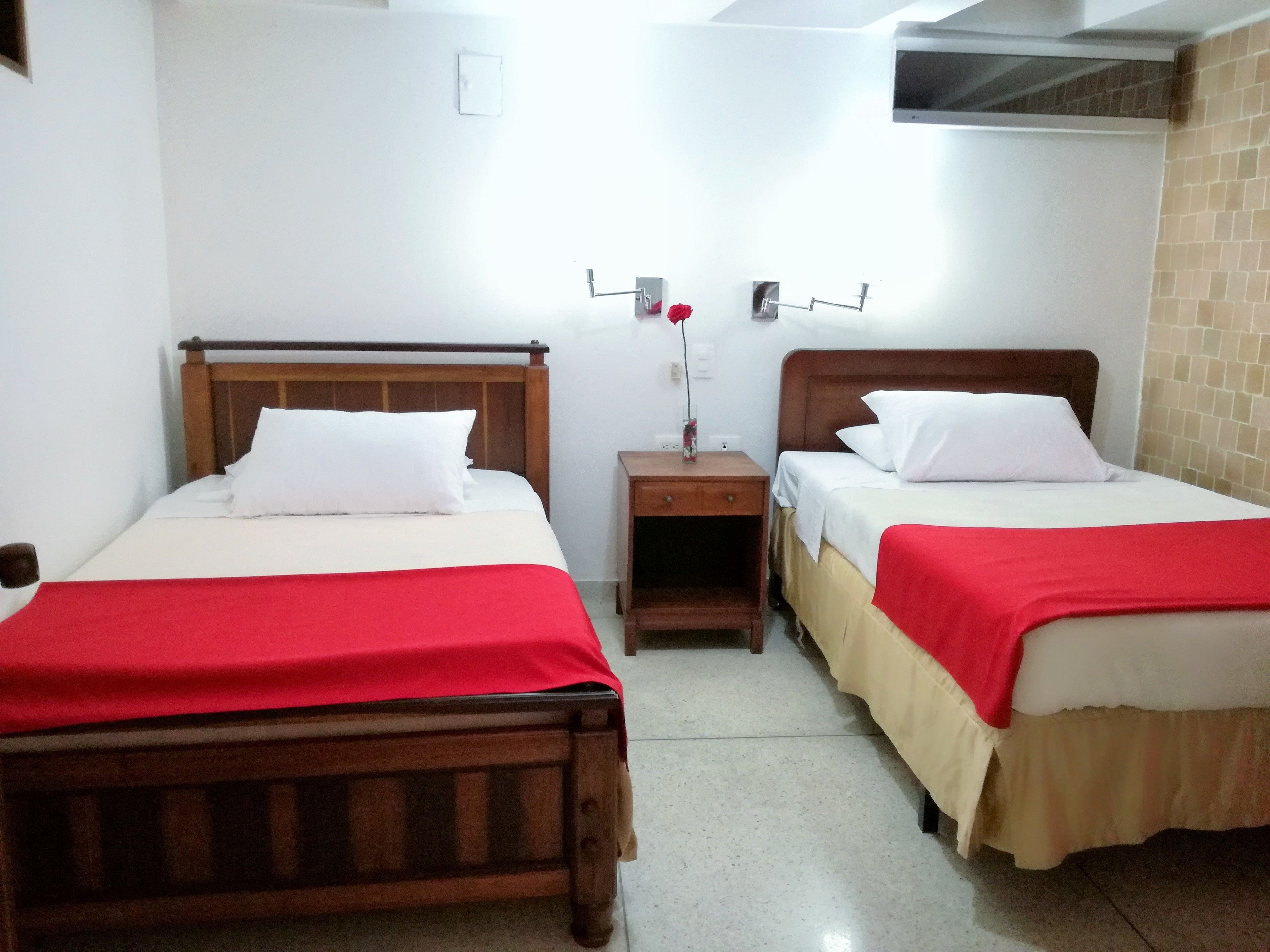 Una camera d'albergo con due letti di legno, ognuno con biancheria bianca e una coperta rossa. Tra i letti si trova un comodino in legno con un vaso di fiori.