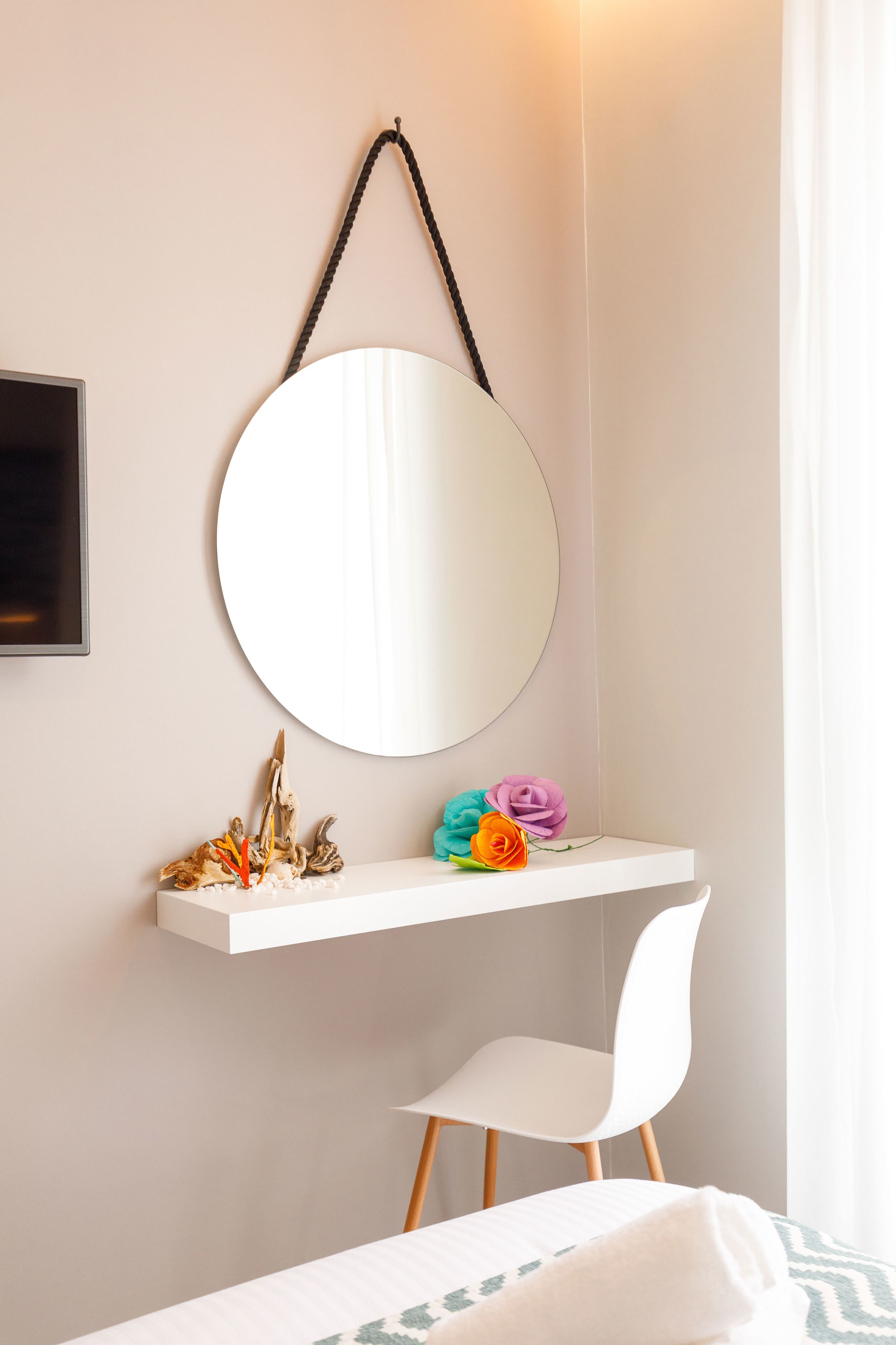 A bedroom with a white bed, a white chair, a bedside table with a mirror, and a small shelf with decorative items.