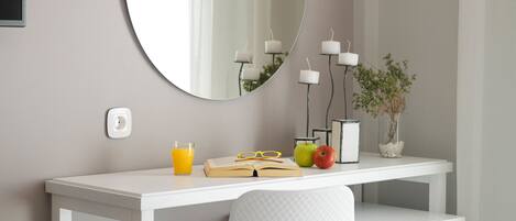 A neatly arranged bedroom with a bed, a bedside table with a lamp, a mirror, and a small desk with a plant and candles.