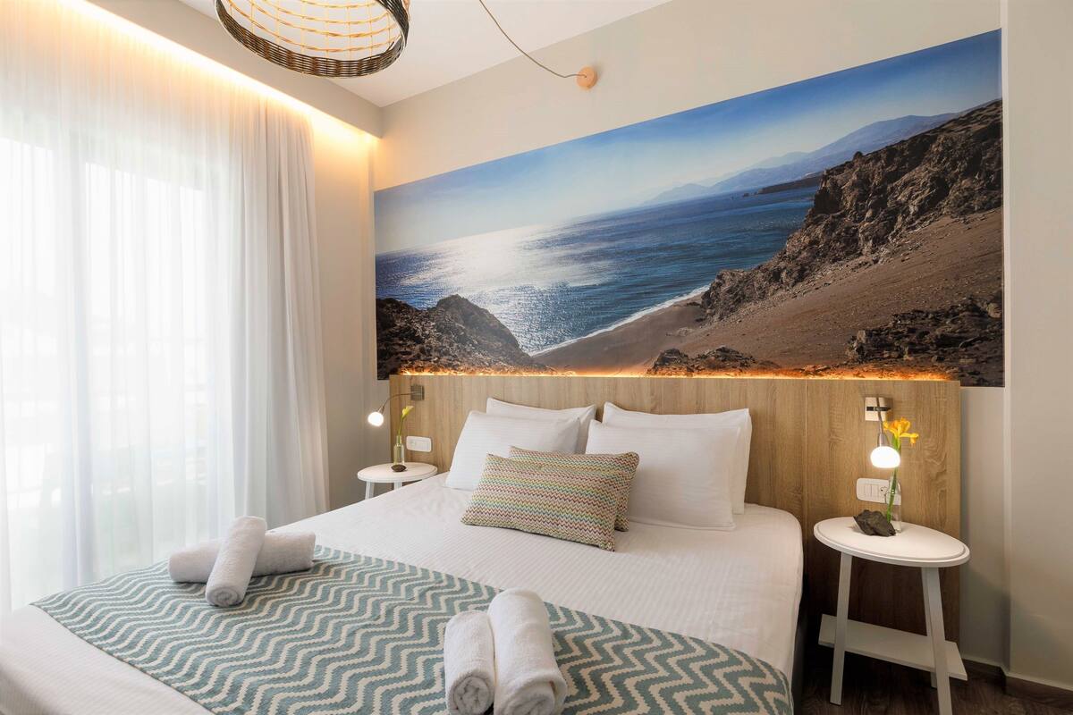 A hotel room with a large bed, a view of the sea, a wooden headboard, and a small bedside table with a lamp.