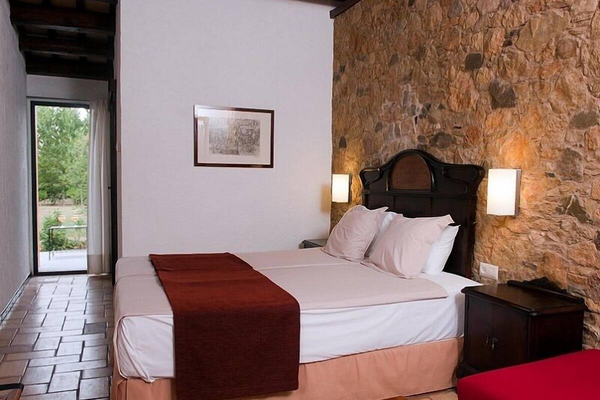 A bedroom with a stone wall, a bed with white linens, a red throw, and two bedside lamps.