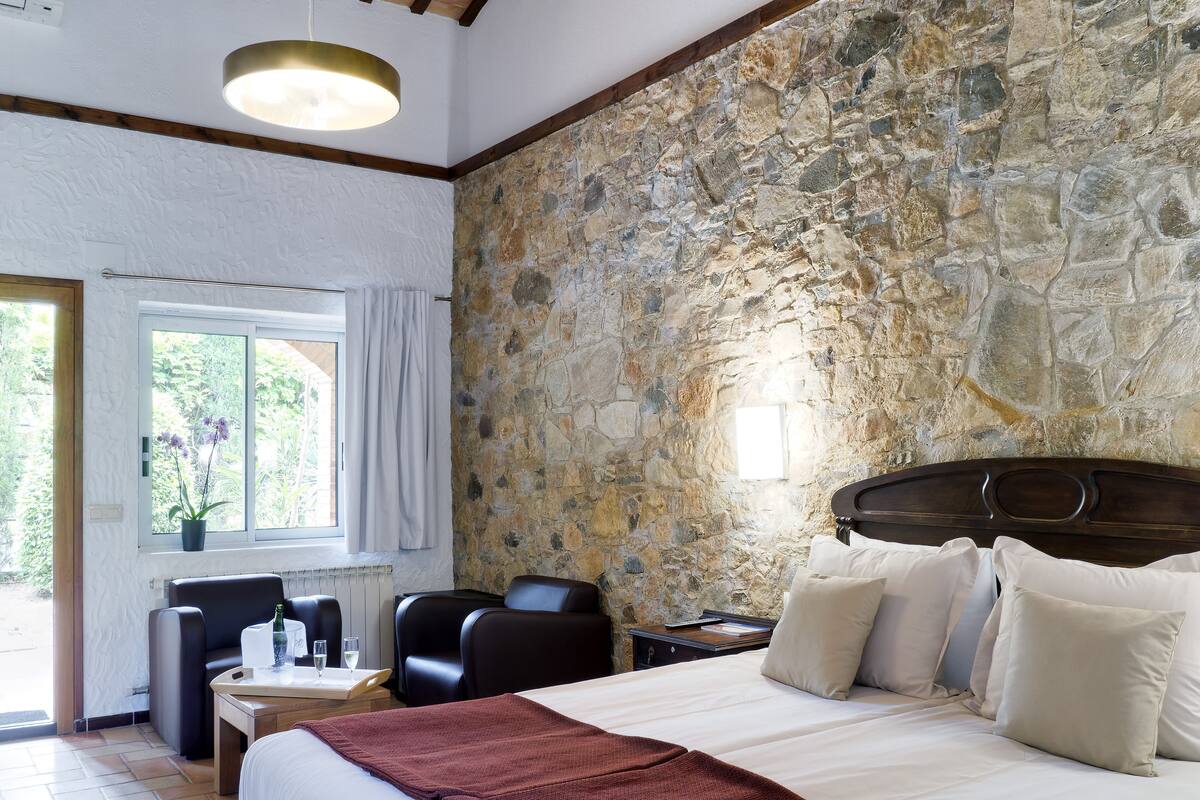 A bedroom with a stone wall, a bed with white linens, a red throw blanket, and a window with curtains.