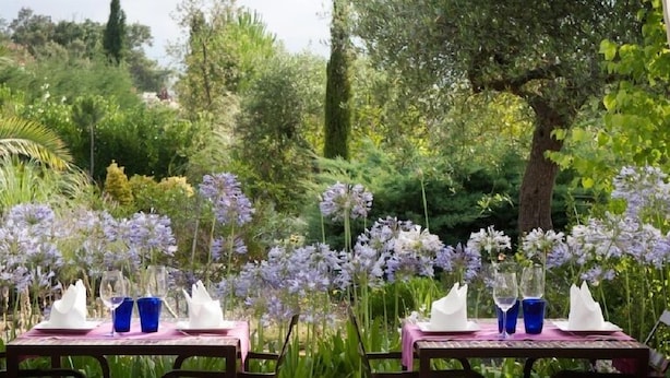 Dos mesas dispuestas al aire libre con servilletas blancas y cristalería azul, rodeadas de flores púrpuras y follaje.