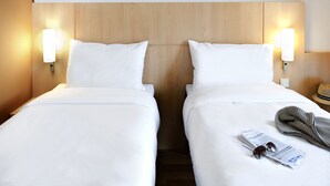 A hotel room with two single beds, each with white bedding and a wooden headboard. There are two wall-mounted light fixtures on either side of the headboard. A hat is visible on the nightstand to the right. - ibis Lyon Est Chaponnay (Chaponnay)