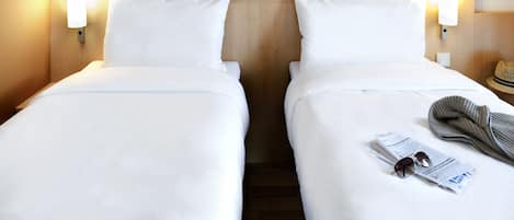 Habitación de hotel con dos camas individuales, cada una con ropa de cama blanca y cabeceros de madera. Hay dos lámparas de pared a cada lado de los cabeceros. Se ve un sombrero sobre la mesita de noche a la derecha.