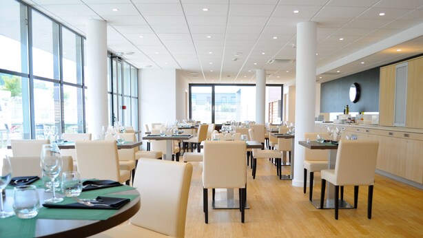 A modern dining area with multiple tables and chairs, large windows, and a kitchen area with cabinets and a clock. - ibis Lyon Est Chaponnay (Chaponnay)