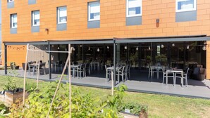 An outdoor dining area with tables and chairs, a covered seating area, and a building with orange and gray brick walls. - ibis Lyon Est Chaponnay (Chaponnay)