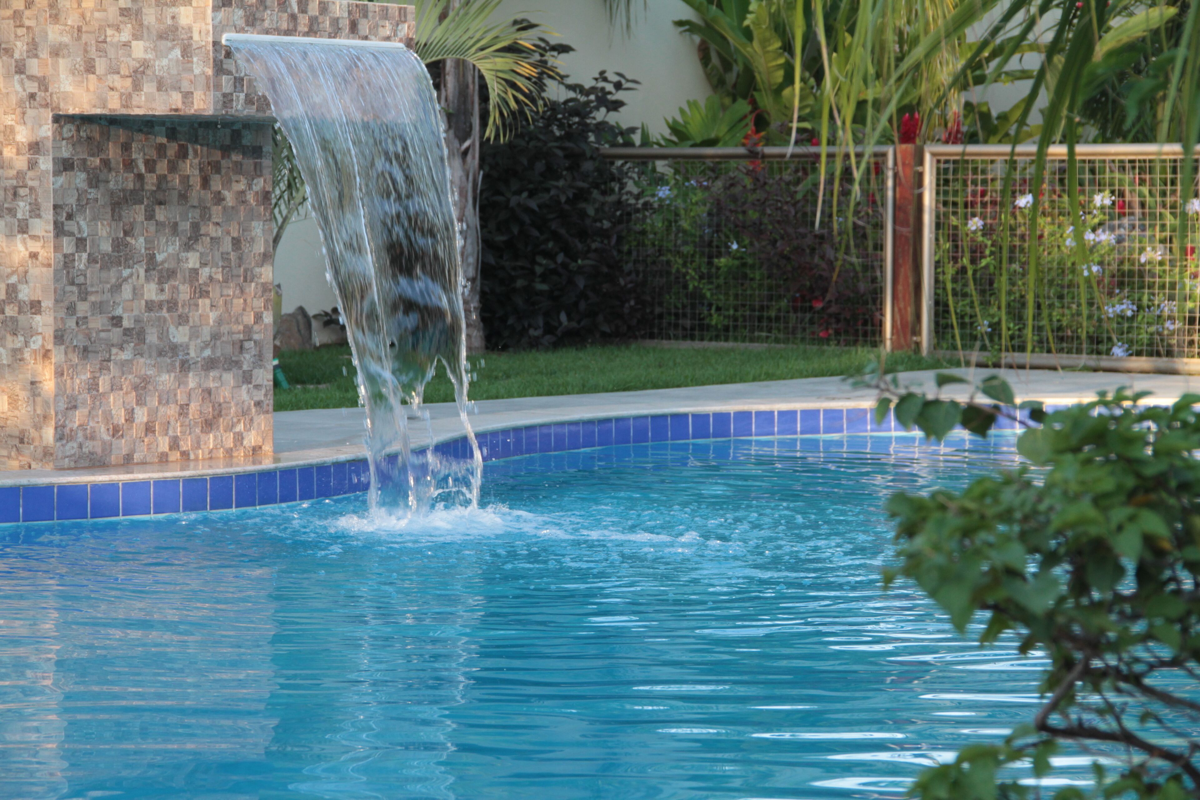 A pool with a waterfall feature, surrounded by a tiled border and greenery.