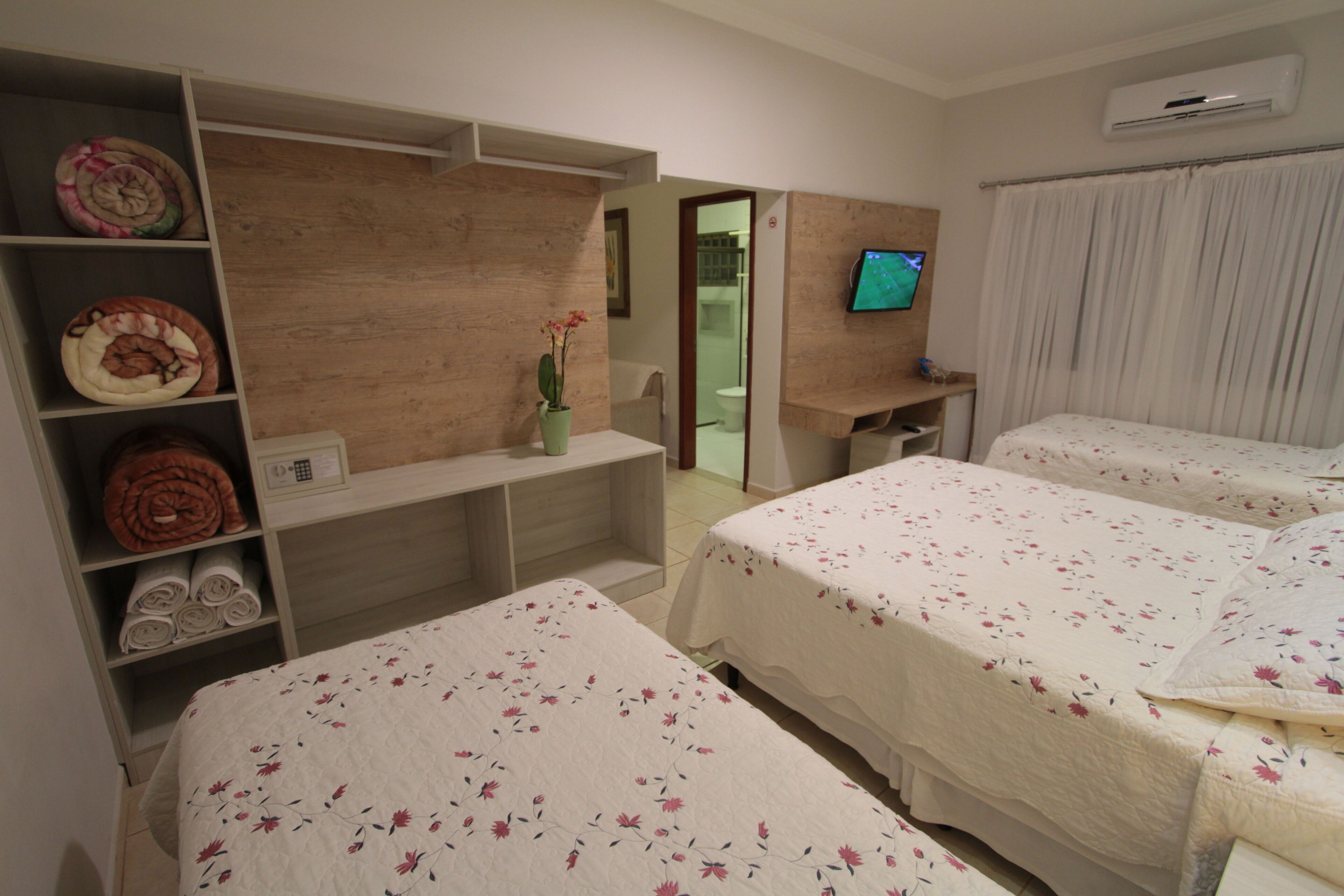 A hotel room with two beds, a wooden shelf with towels, a small table with a vase, and a TV on the wall.