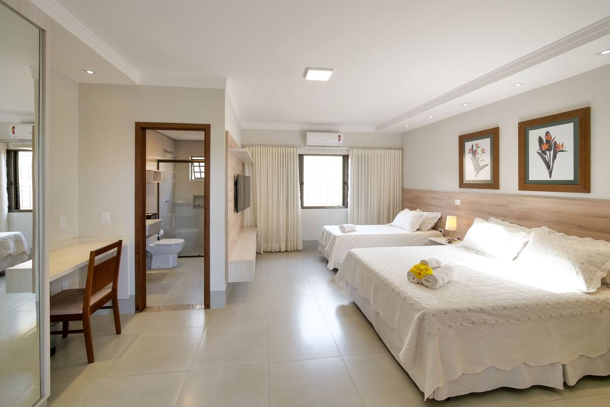 A hotel room with two beds, a desk, a chair, a bathroom, and a large window.
