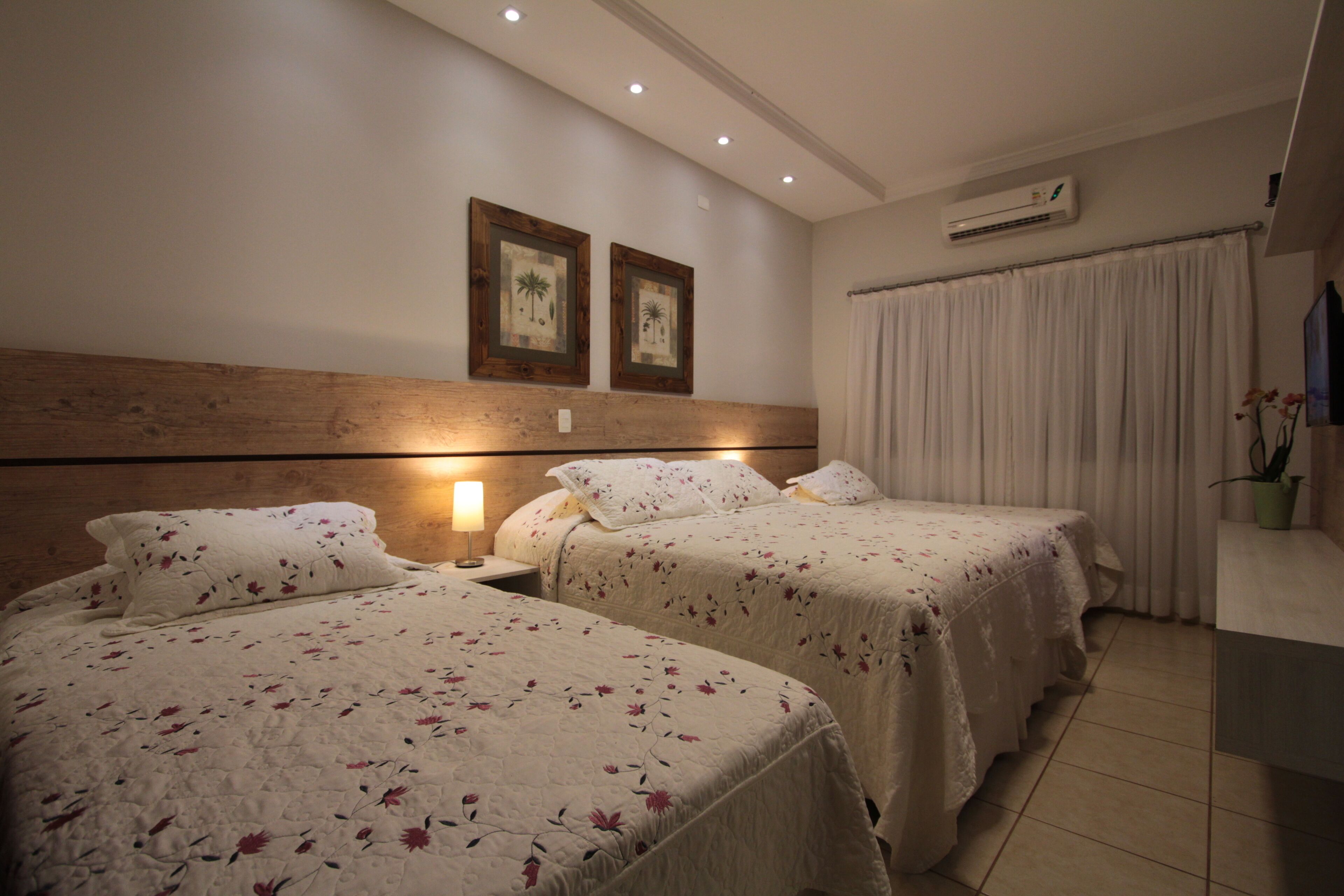 Quarto de hotel com duas camas, cabeceira de madeira, mesa de cabeceira, luminária, planta em vaso e aparelho de ar condicionado.