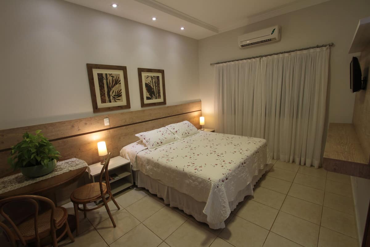 A hotel room with a bed, a desk with a chair, a potted plant, and two framed pictures on the wall.