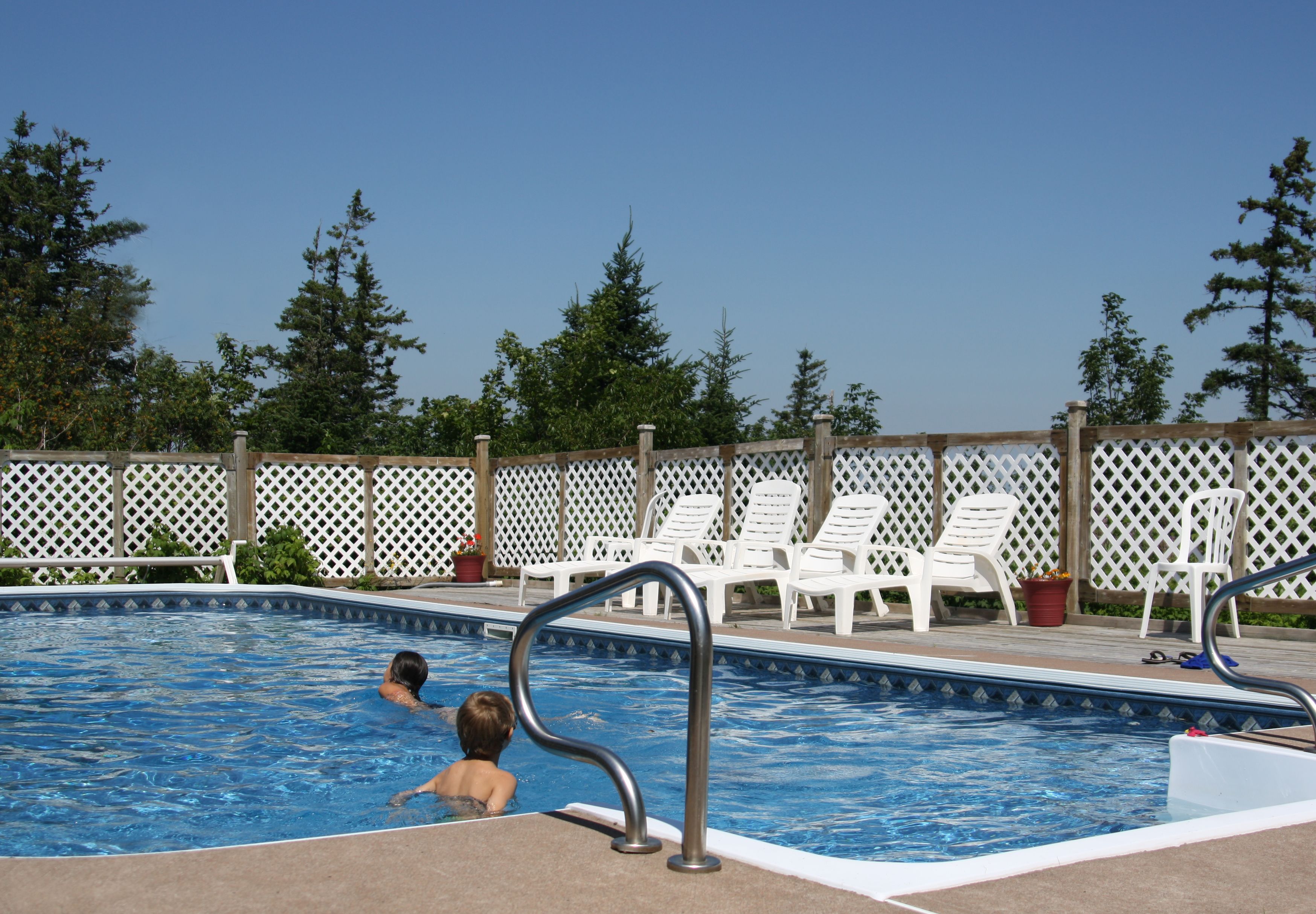 A swimming pool with a metal ladder, surrounded by white lounge chairs and a wooden fence.