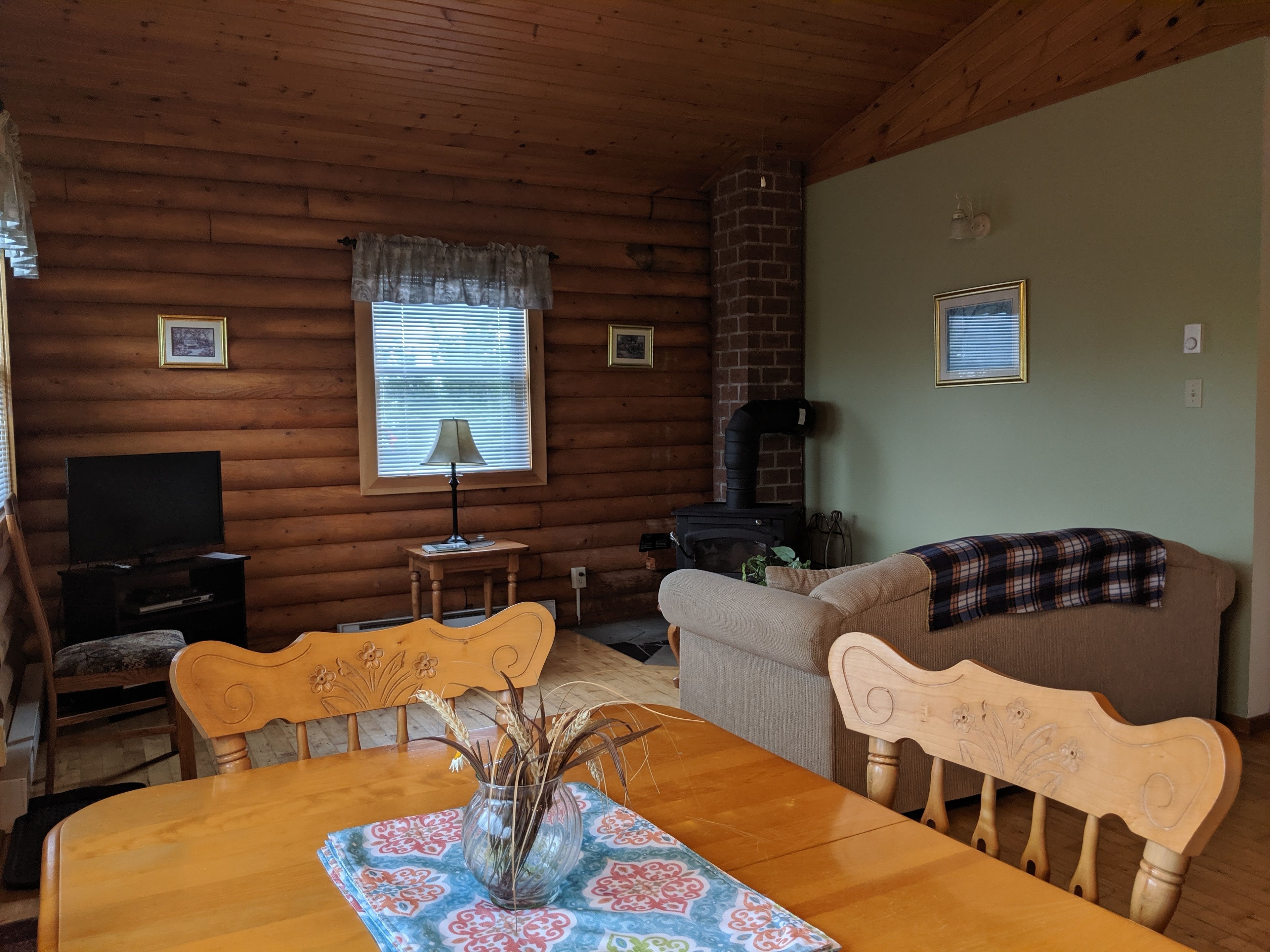 A cozy log cabin interior with a dining table, chairs, a sofa, a television, a fireplace, and framed pictures on the walls.