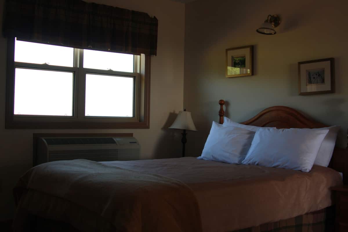 Ein Hotelzimmer mit großem Fenster, einem Bett mit weißen Laken, einem Holz-Kopfschild, einer Nachttischlampe und einer Wand-Klimaanlage.