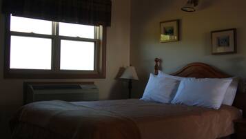 A hotel room with a large window, a bed with white linens, a wooden headboard, a bedside lamp, and a wall-mounted air conditioner.