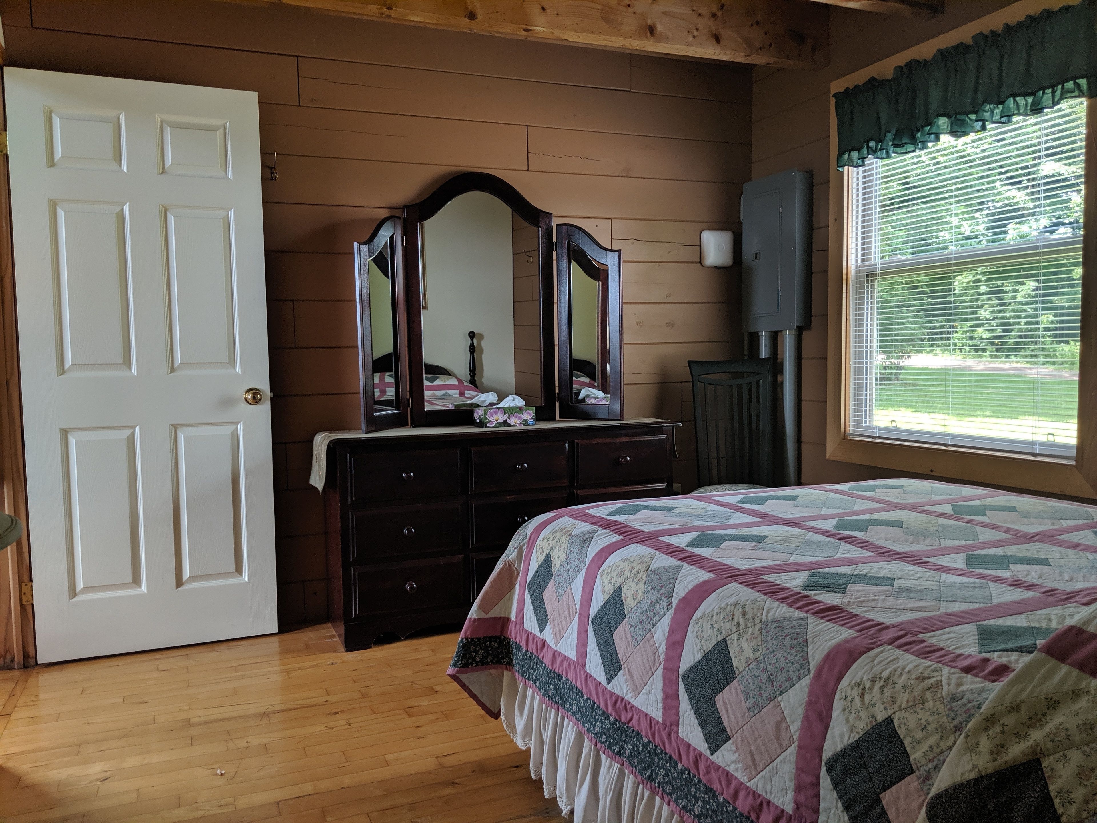 A bedroom with a wooden floor, a bed with a quilt, a dresser with a mirror, a window with blinds, and a door.