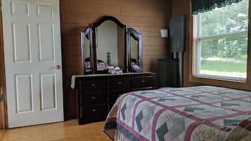 A bedroom with a wooden floor, a bed with a quilt, a dresser with a mirror, a window with blinds, and a door.