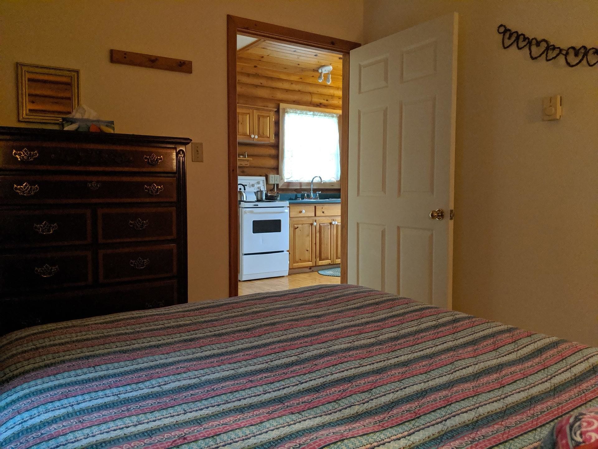 A bedroom with a bed, a dresser, and a view into a kitchen area with wooden cabinets and a stove.