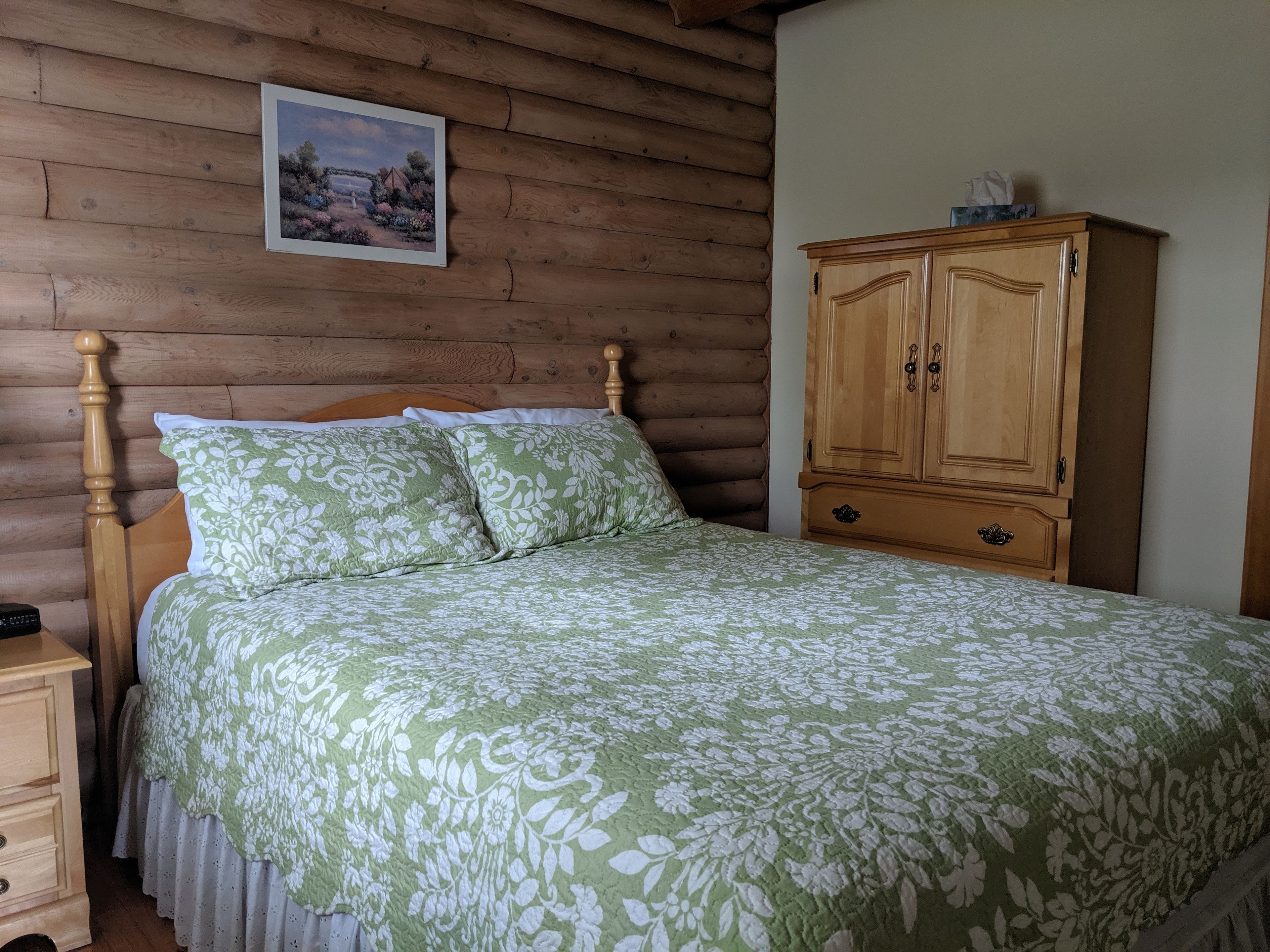A wooden log cabin-style bedroom with a bed, a wooden nightstand, a wooden wardrobe, and a framed picture on the wall.