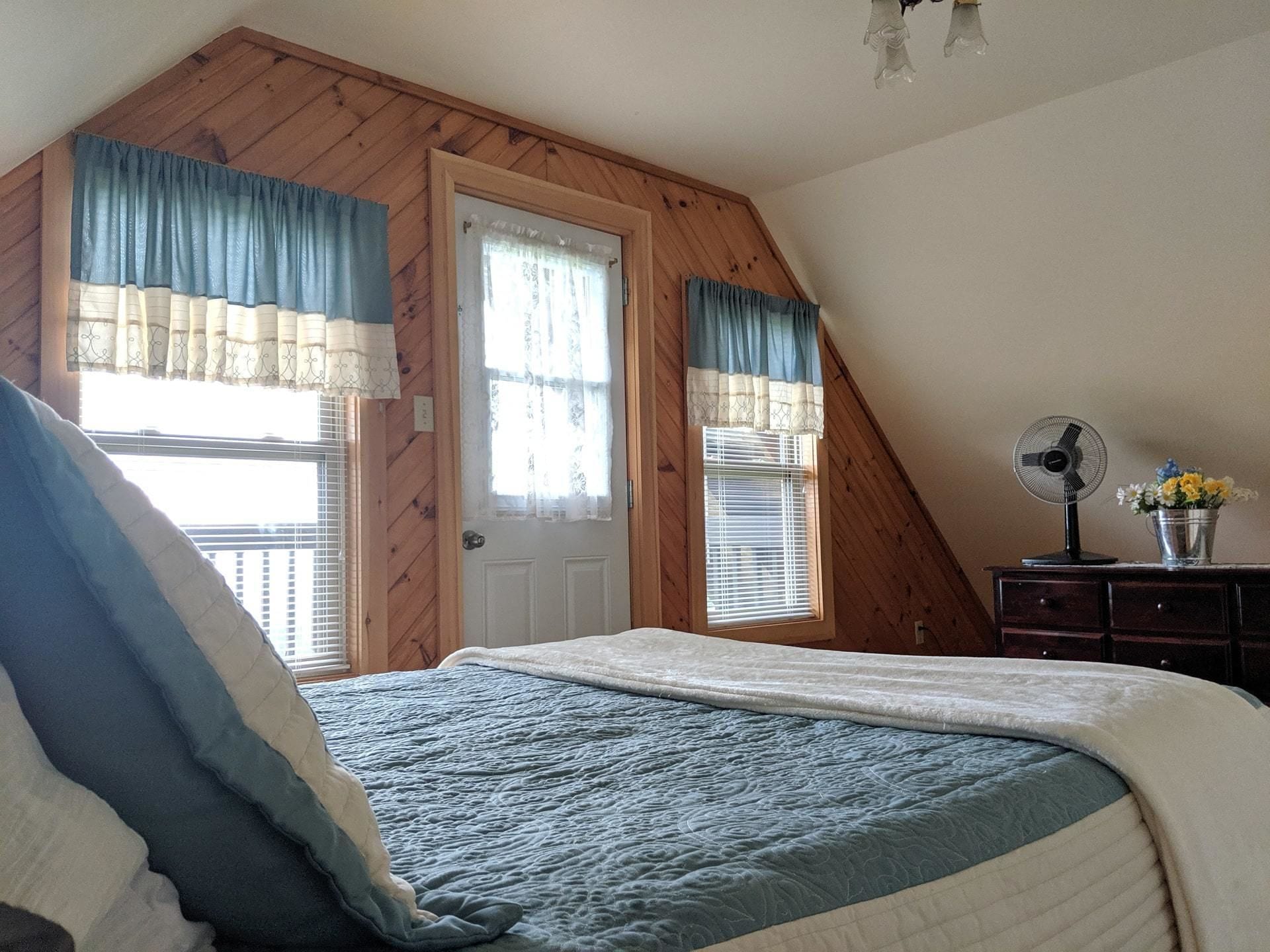 A bedroom with a sloped ceiling, a bed with a blue and white quilt, a fan, and a dresser with a vase of flowers.