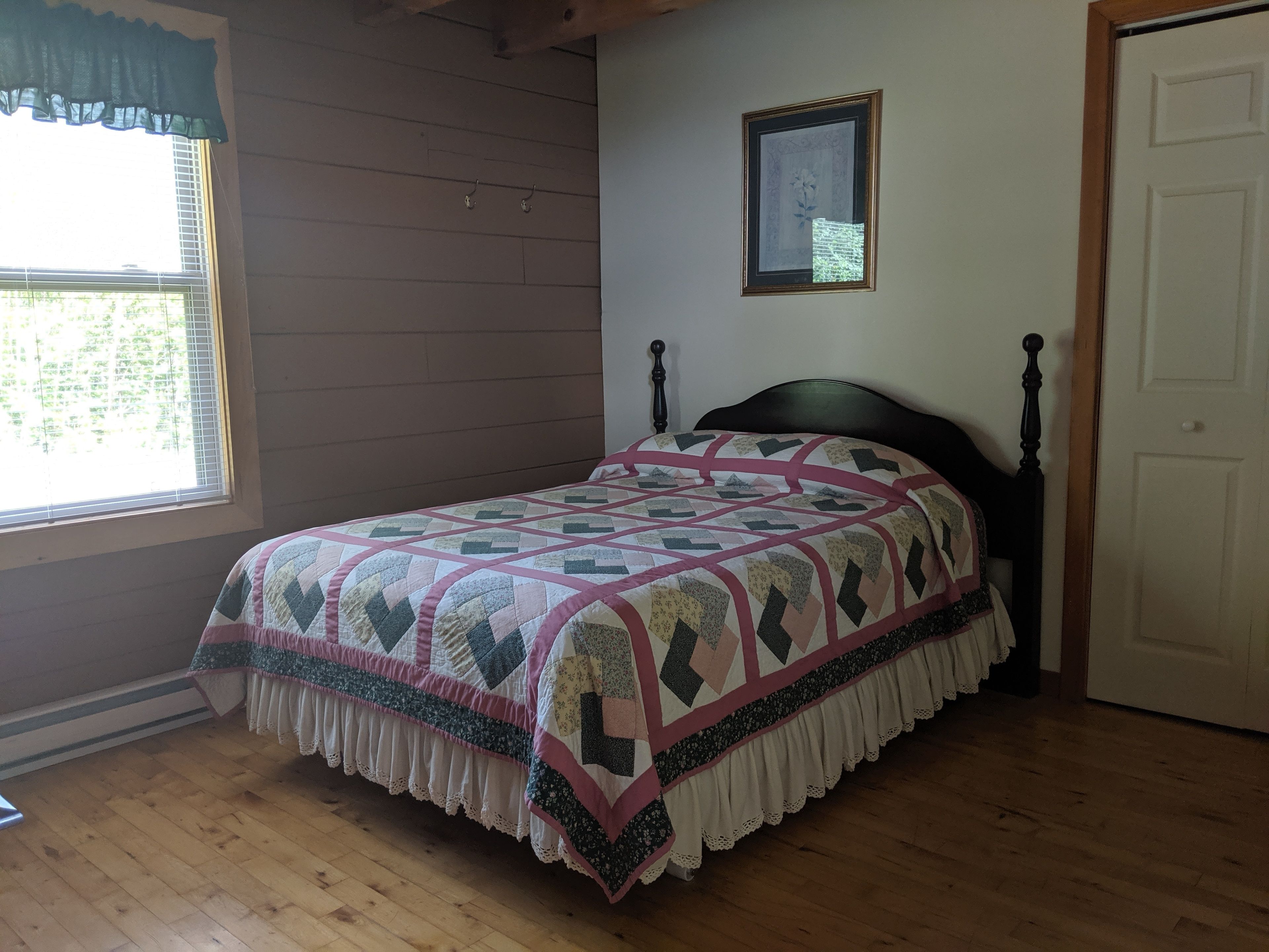 Quarto com piso de madeira, cama com colcha estampada, janela com persianas e uma porta.