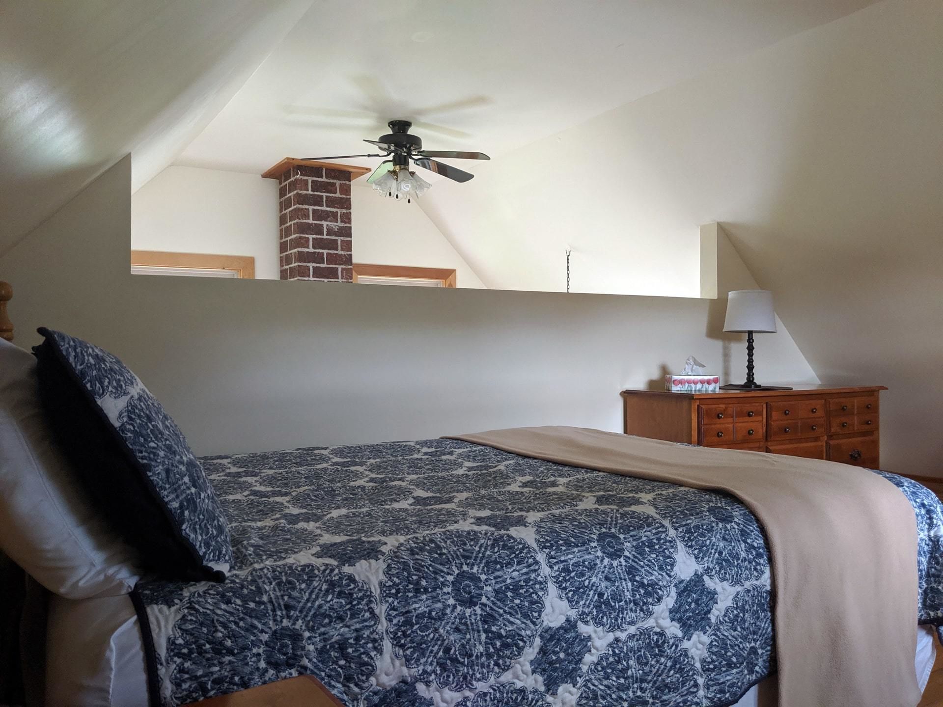 Quarto com cama, mesa de cabeceira, cômoda, ventoinha no teto e lareira de tijolos.