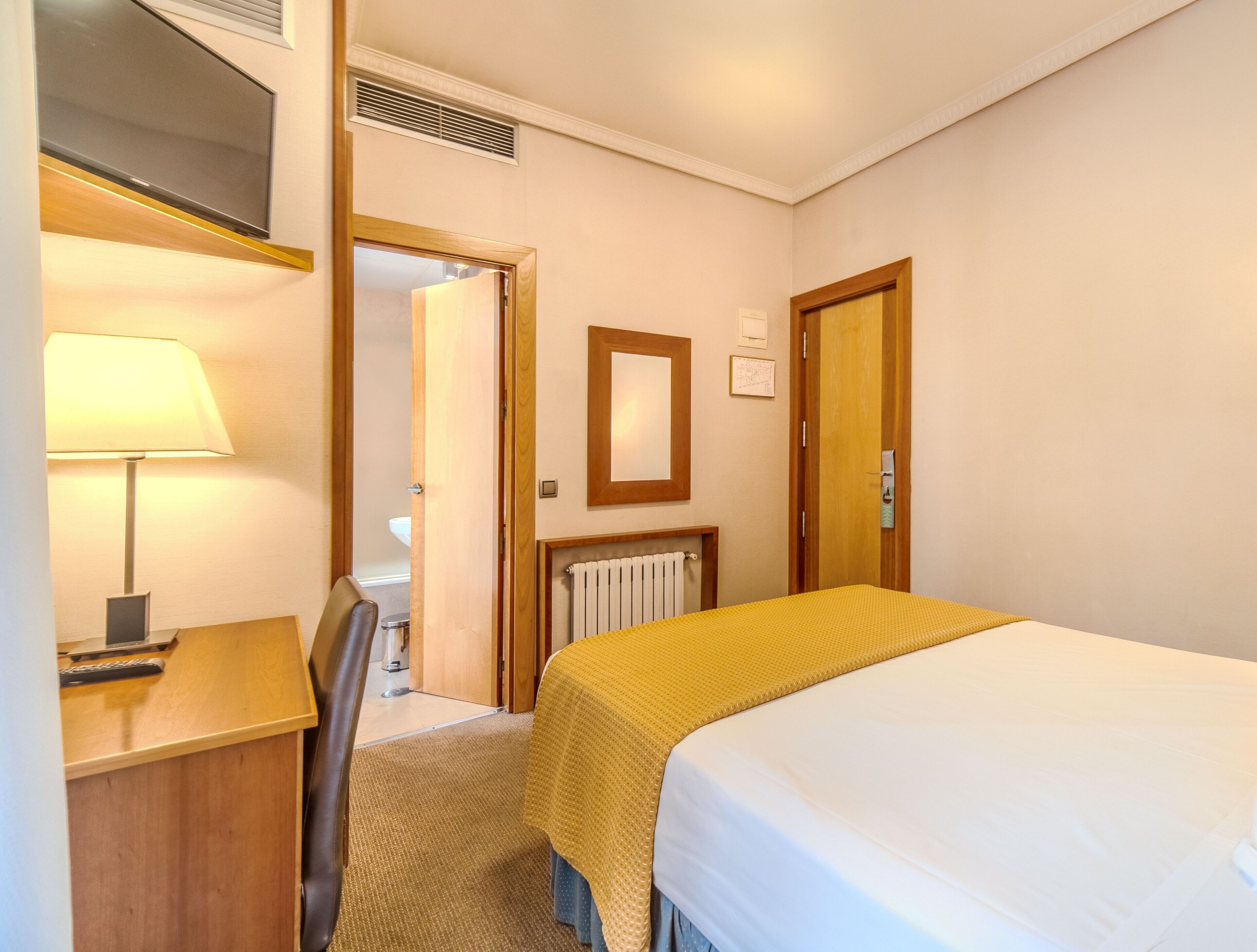 Habitación de hotel con cama, escritorio, silla, lámpara, radiador y espejo en la pared.