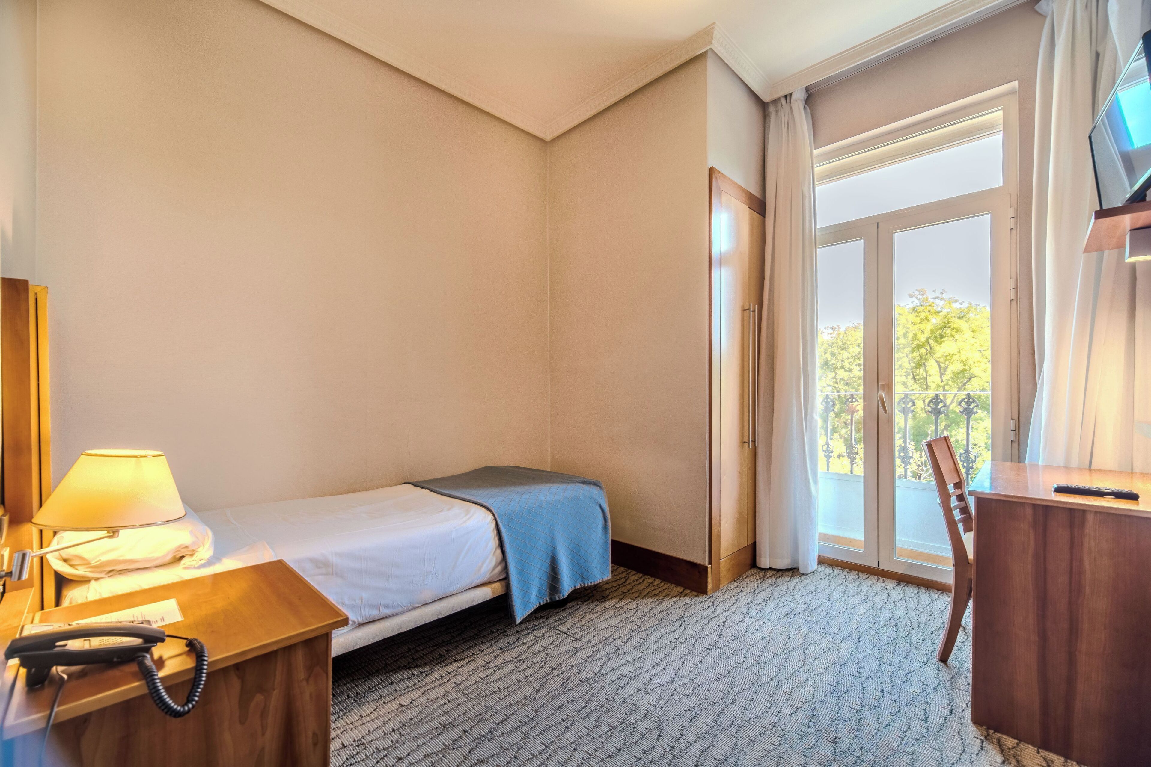 Habitación de hotel con cama, escritorio con lámpara, teléfono y vista al exterior a través de una ventana.