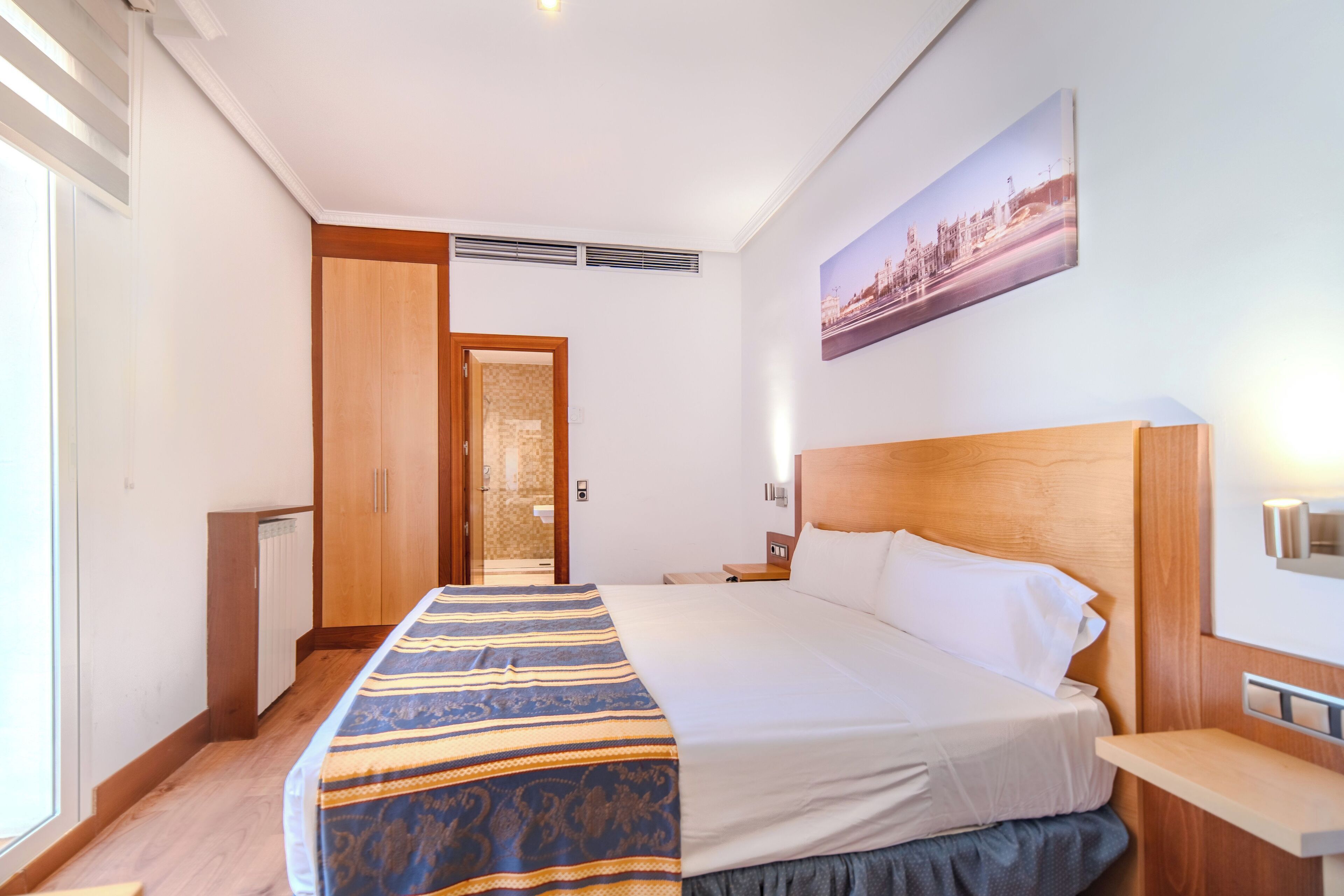 A hotel room with a large bed, wooden headboard, a nightstand, a bedside table, a window, and a door leading to a bathroom.