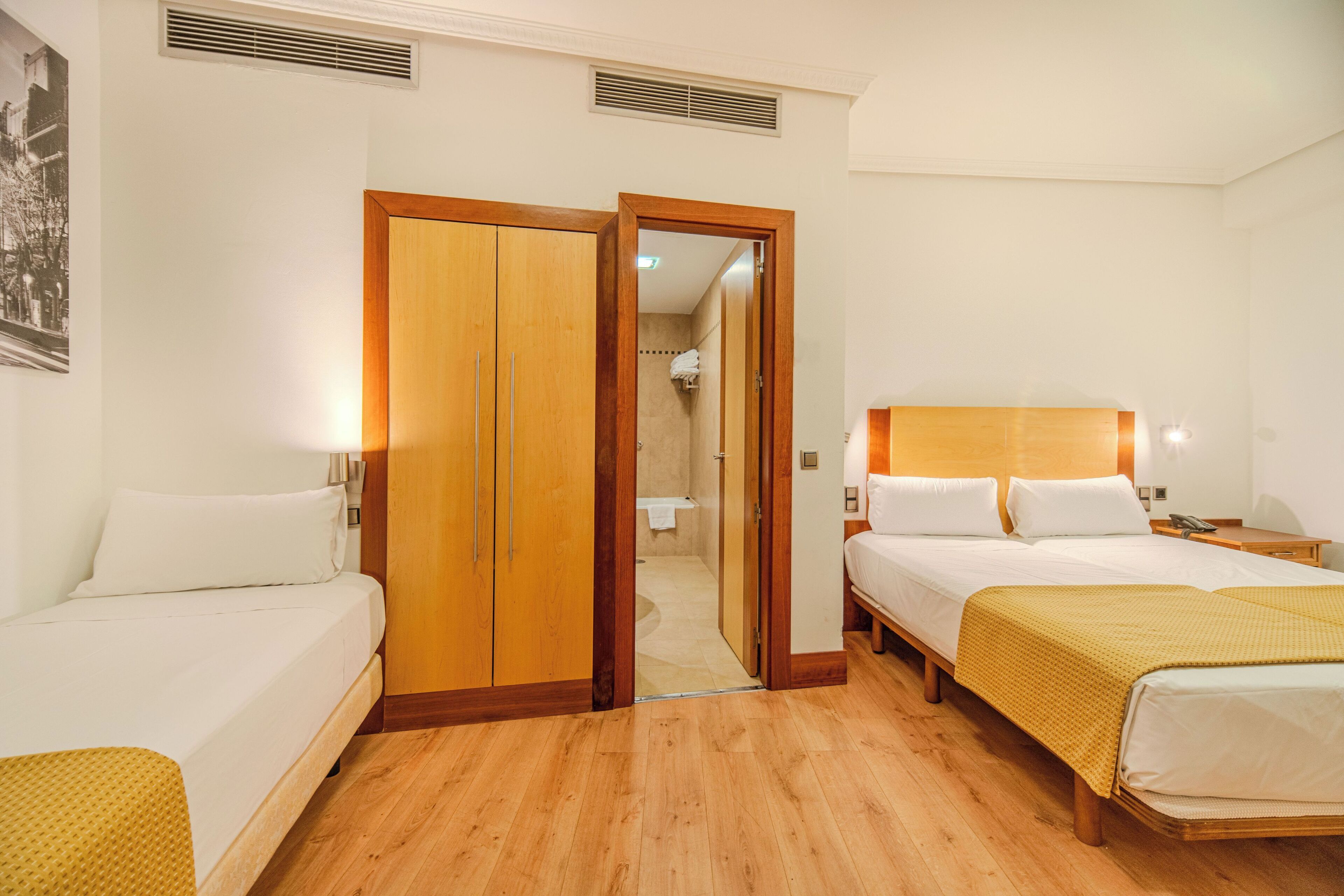Een hotelkamer met twee bedden, houten vloeren, een kledingkast en een badkamer zichtbaar door een geopende deur.