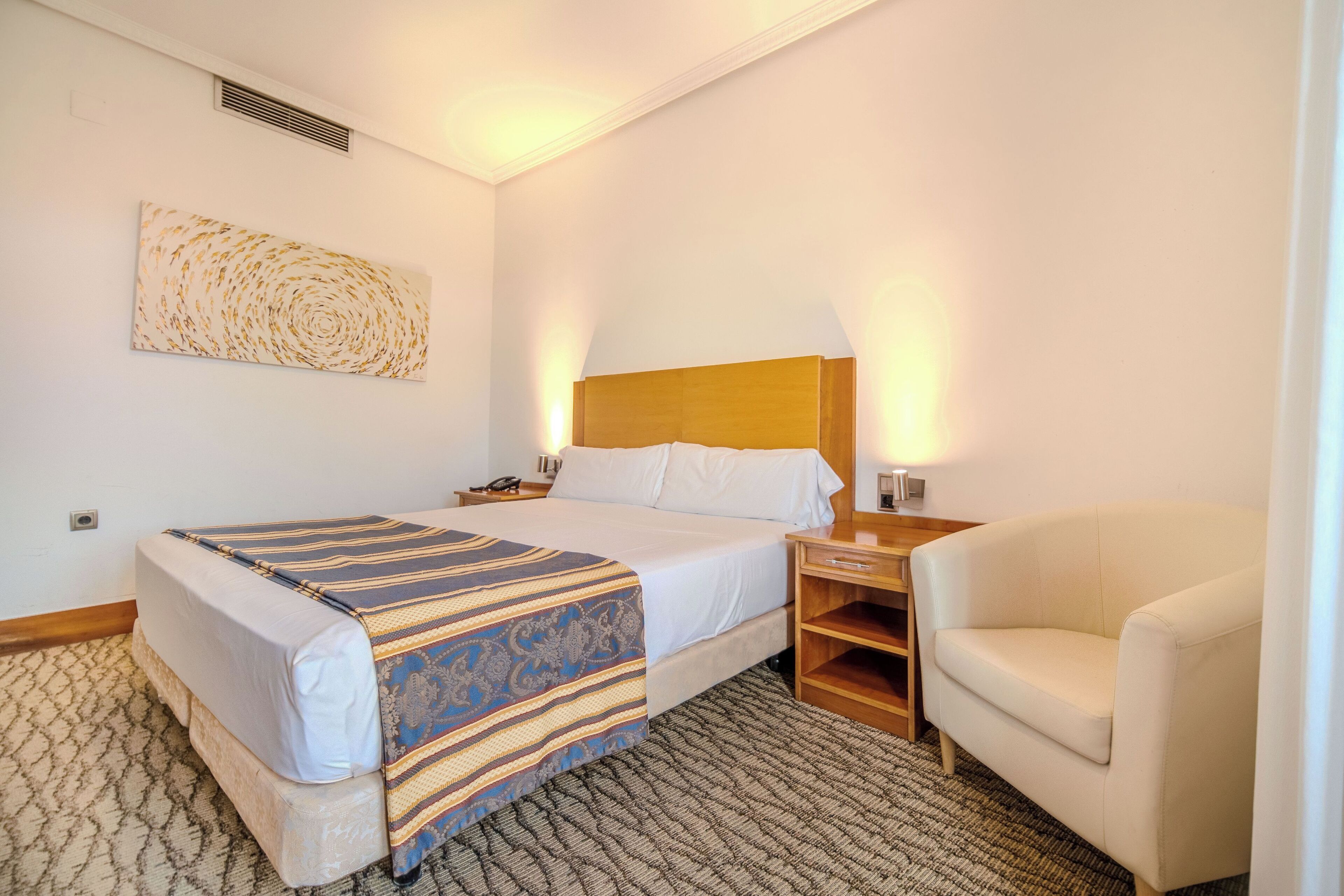 A hotel room with a bed, a nightstand, a lamp, a chair, and a wall art piece.