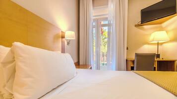 Una camera d'albergo con un letto, una scrivania, una sedia, una TV a muro e una finestra con tende.