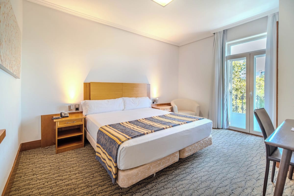 A hotel room with a large bed, a bedside table with a lamp, a chair, a desk, and a view of the outdoors through a window.