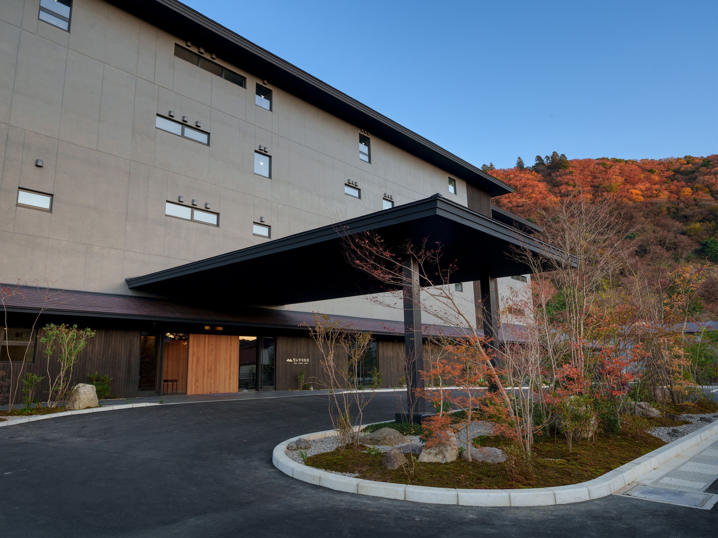A modern building with a covered entrance, a landscaped area with rocks and plants, and a view of autumn-colored trees in the background.