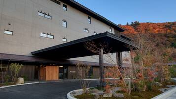 A modern building with a covered entrance, a landscaped area with rocks and plants, and a view of autumn-colored trees in the background.