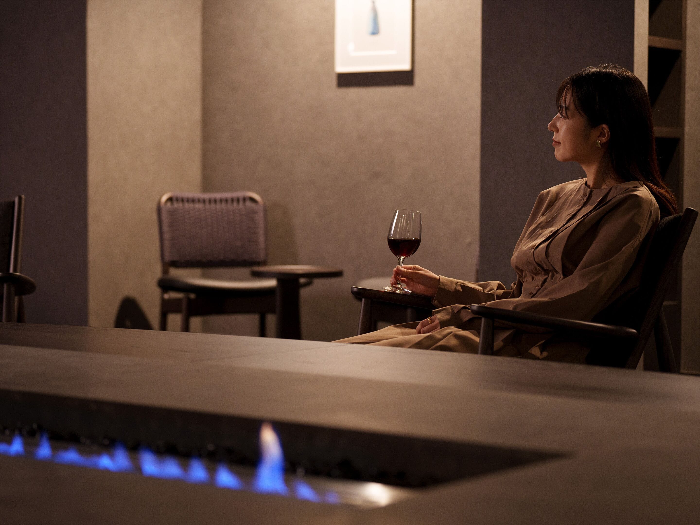 A person seated at a table with a glass of red wine, a chair, and a small table in a dimly lit room with a fireplace.