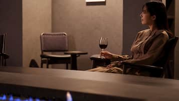 A person seated at a table with a glass of red wine, a chair, and a small table in a dimly lit room with a fireplace.