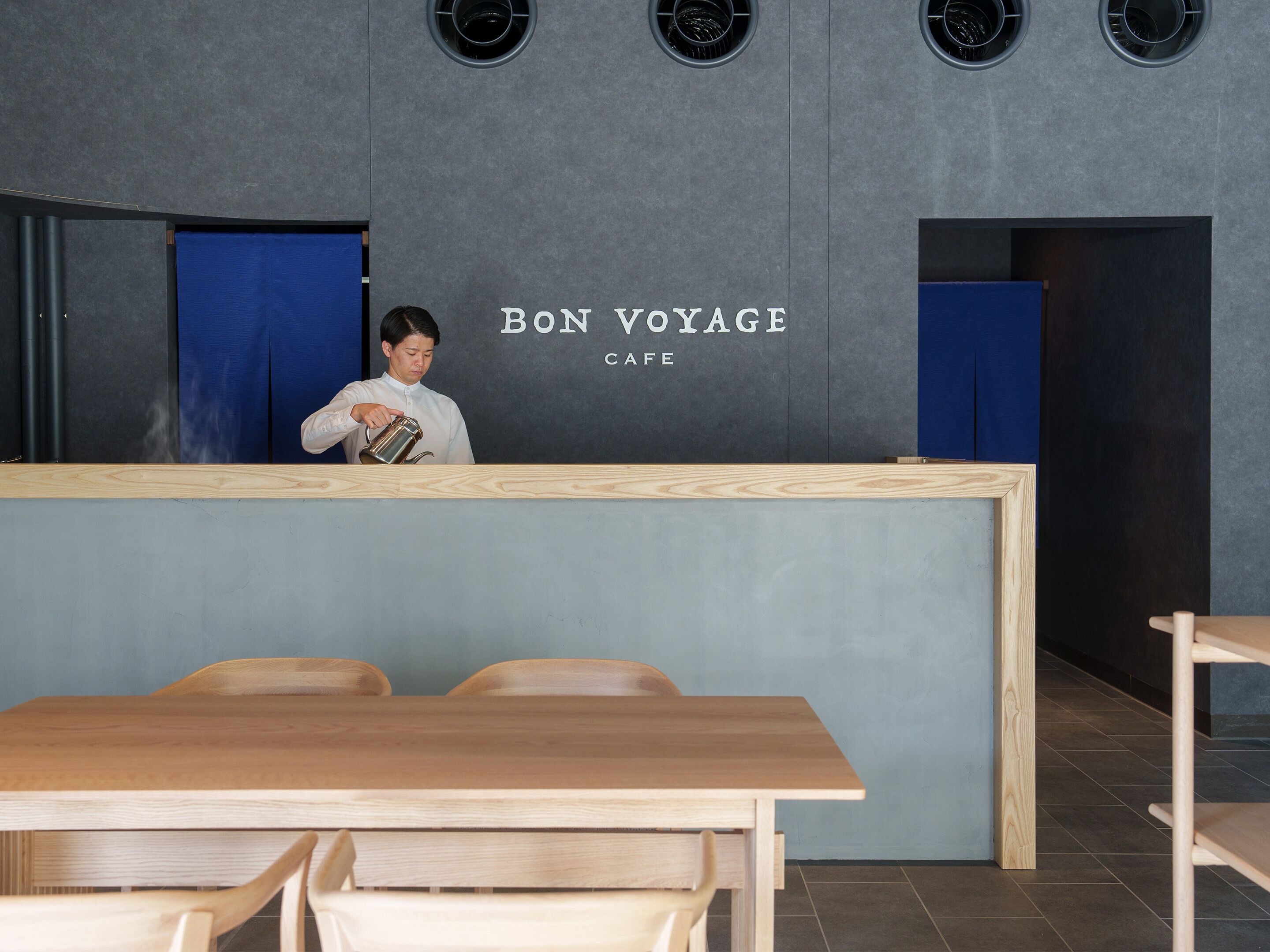 Un mostrador de recepción con un letrero que dice "BON VOYAGE CAFE" y una persona sirviendo una bebida. Se ven sillas de madera y una mesa en primer plano.