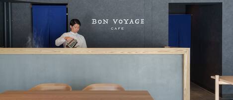 Un mostrador de recepción con un letrero que dice "BON VOYAGE CAFE" y una persona sirviendo una bebida. Se ven sillas de madera y una mesa en primer plano.