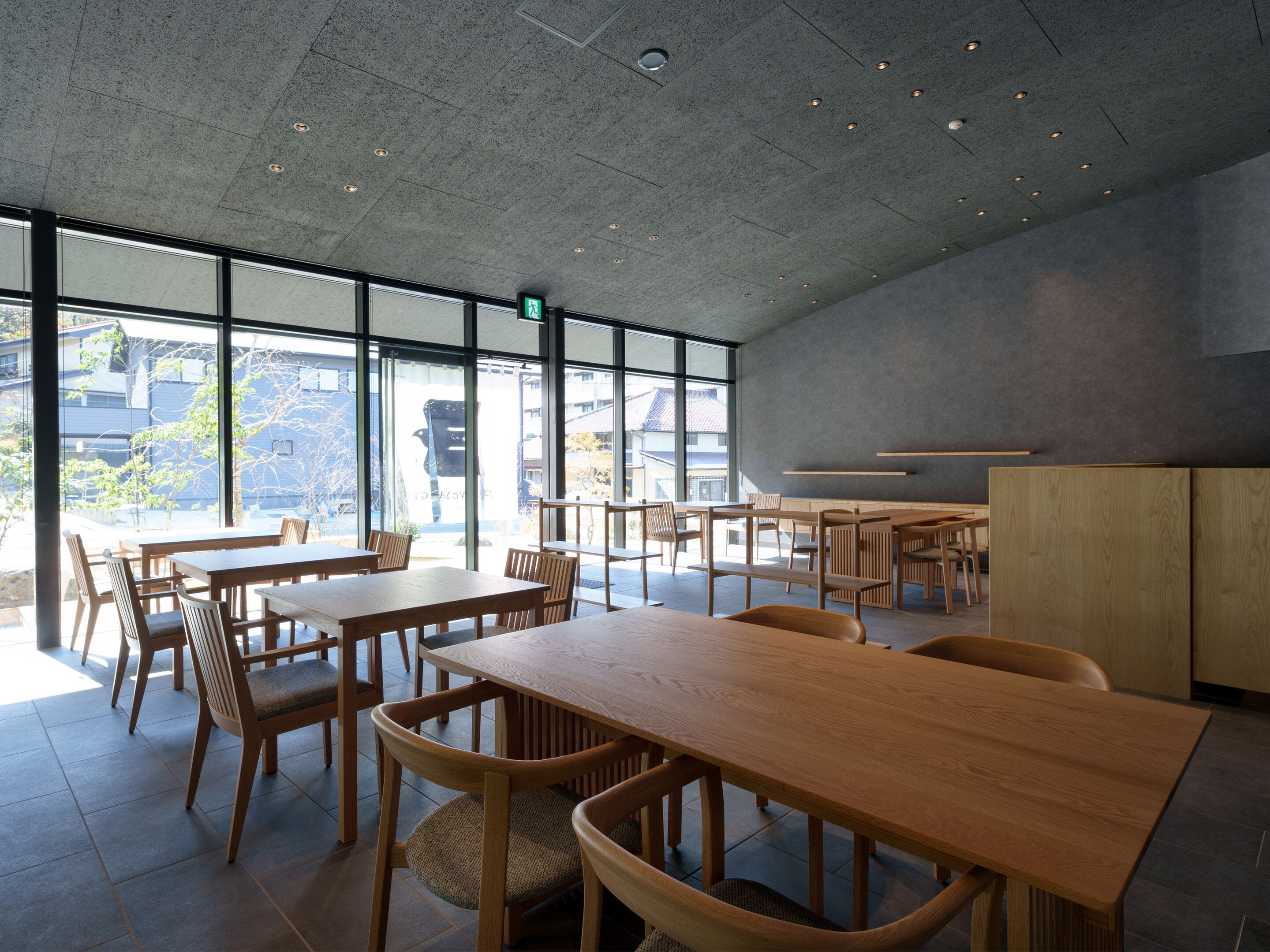 A spacious indoor area with multiple wooden tables and chairs, large windows allowing natural light, and a modern ceiling design.