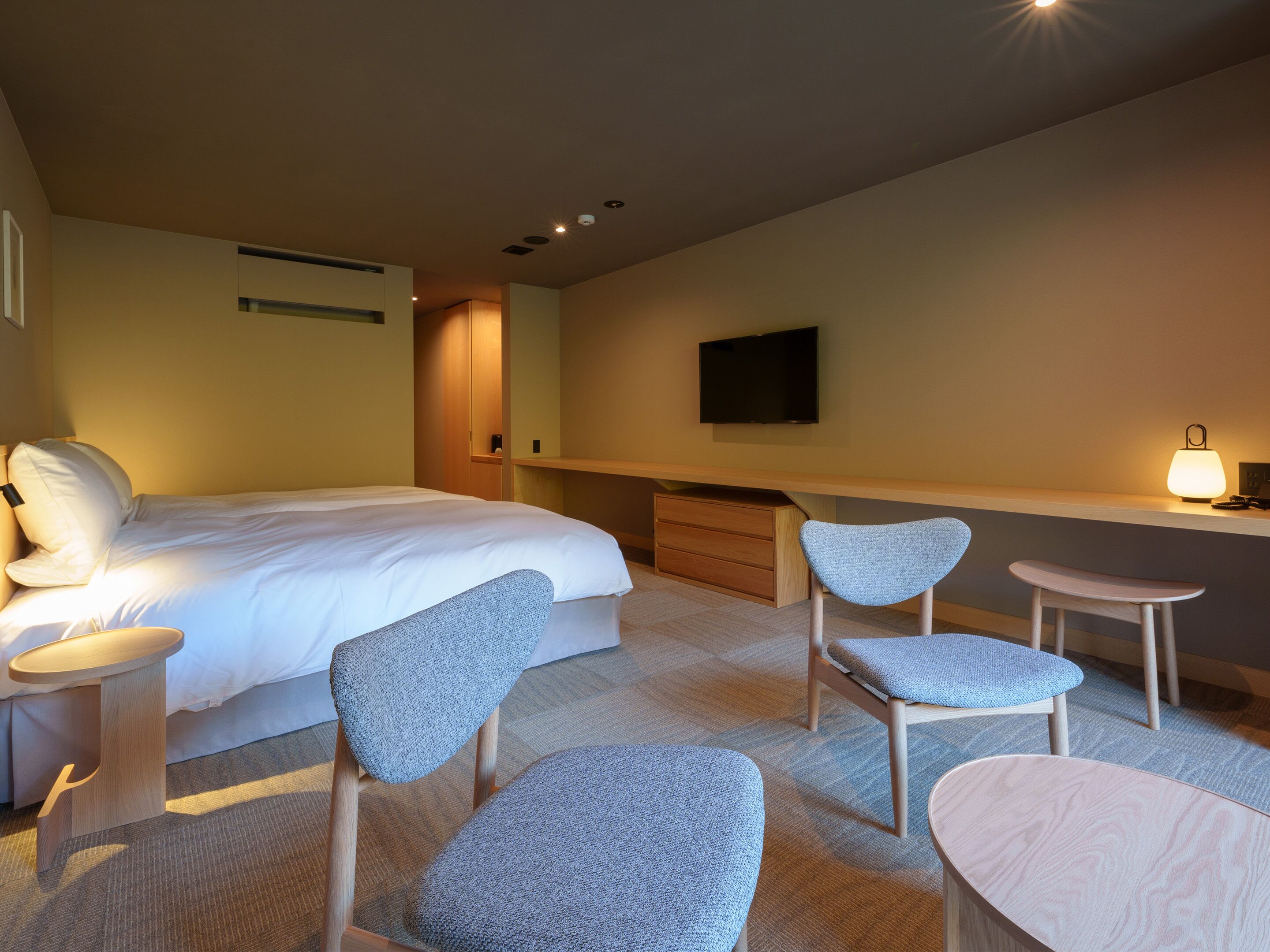 A hotel room with a bed, two chairs, a desk, a TV, and a bedside lamp.