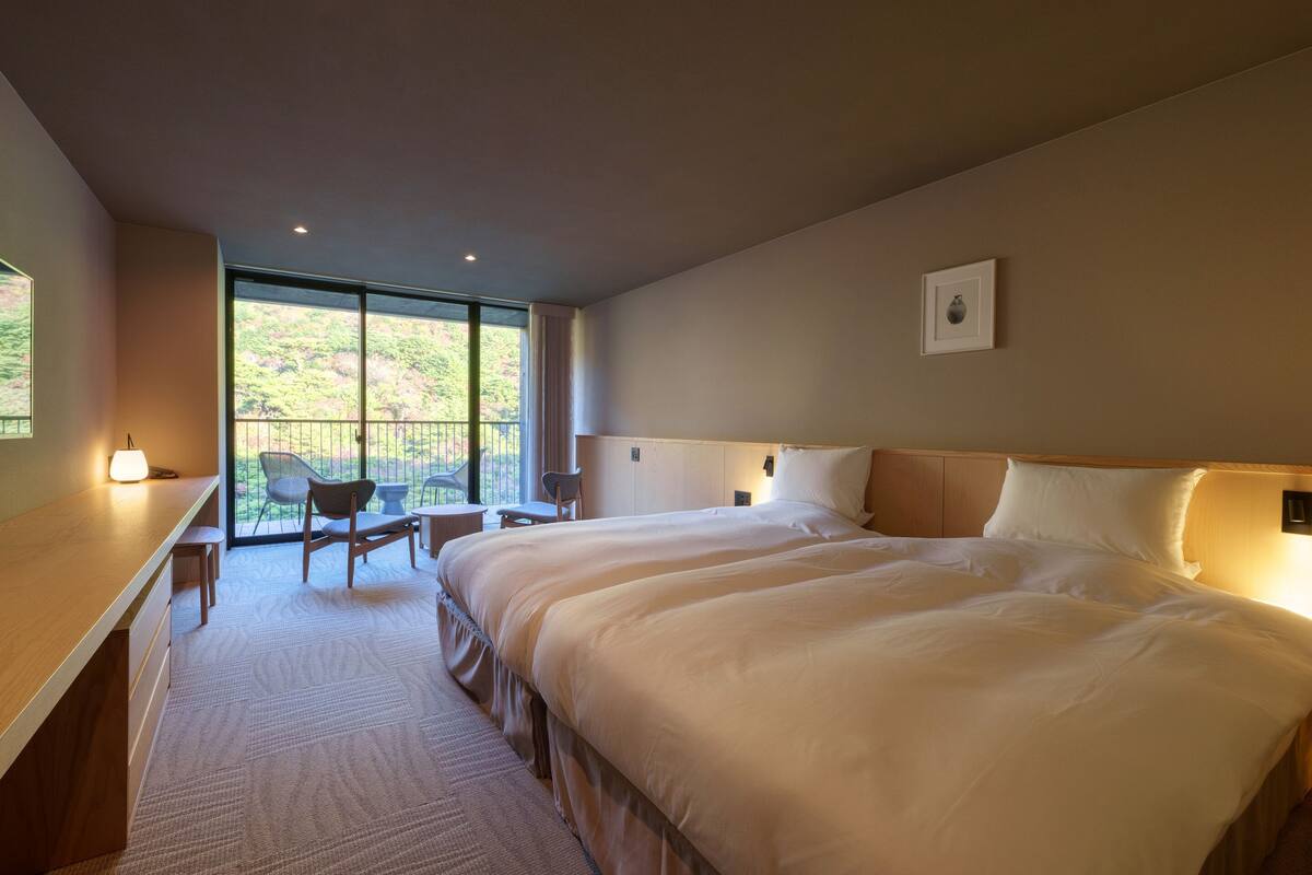 A modern hotel room with a large bed, a desk, and a balcony with chairs.