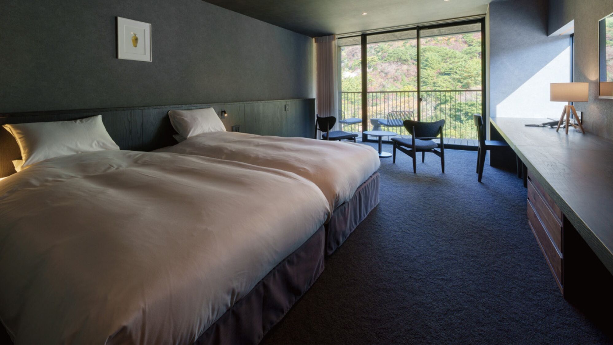 A bedroom with a large bed, a desk, and a chair, with a view of greenery outside.